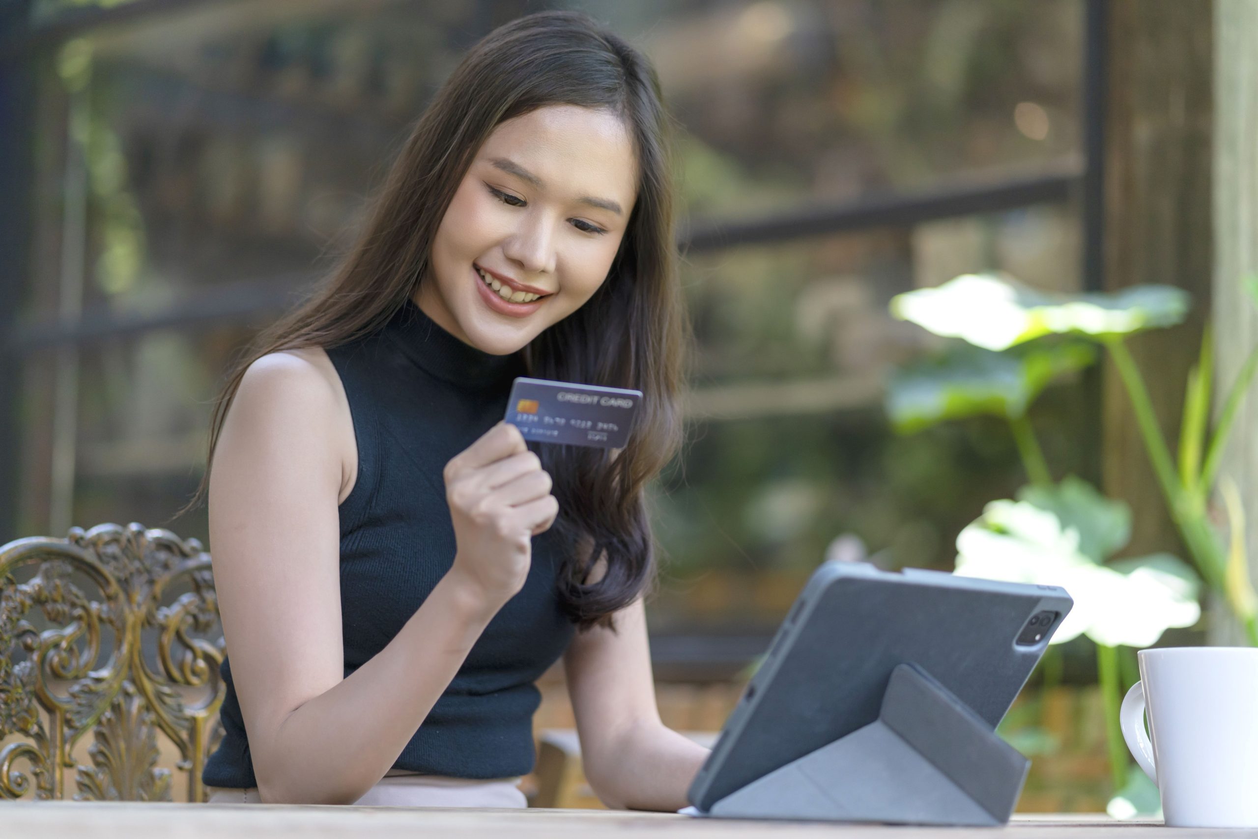 A imagem é de uma mulher asiática jovem, branca, de cabelos castanhos escuros e blusa preta sentada em uma mesa olhando um cartão de crédito azul escuro e sorrindo. Em frente a ela há um tablet preto apoiado na mesa.