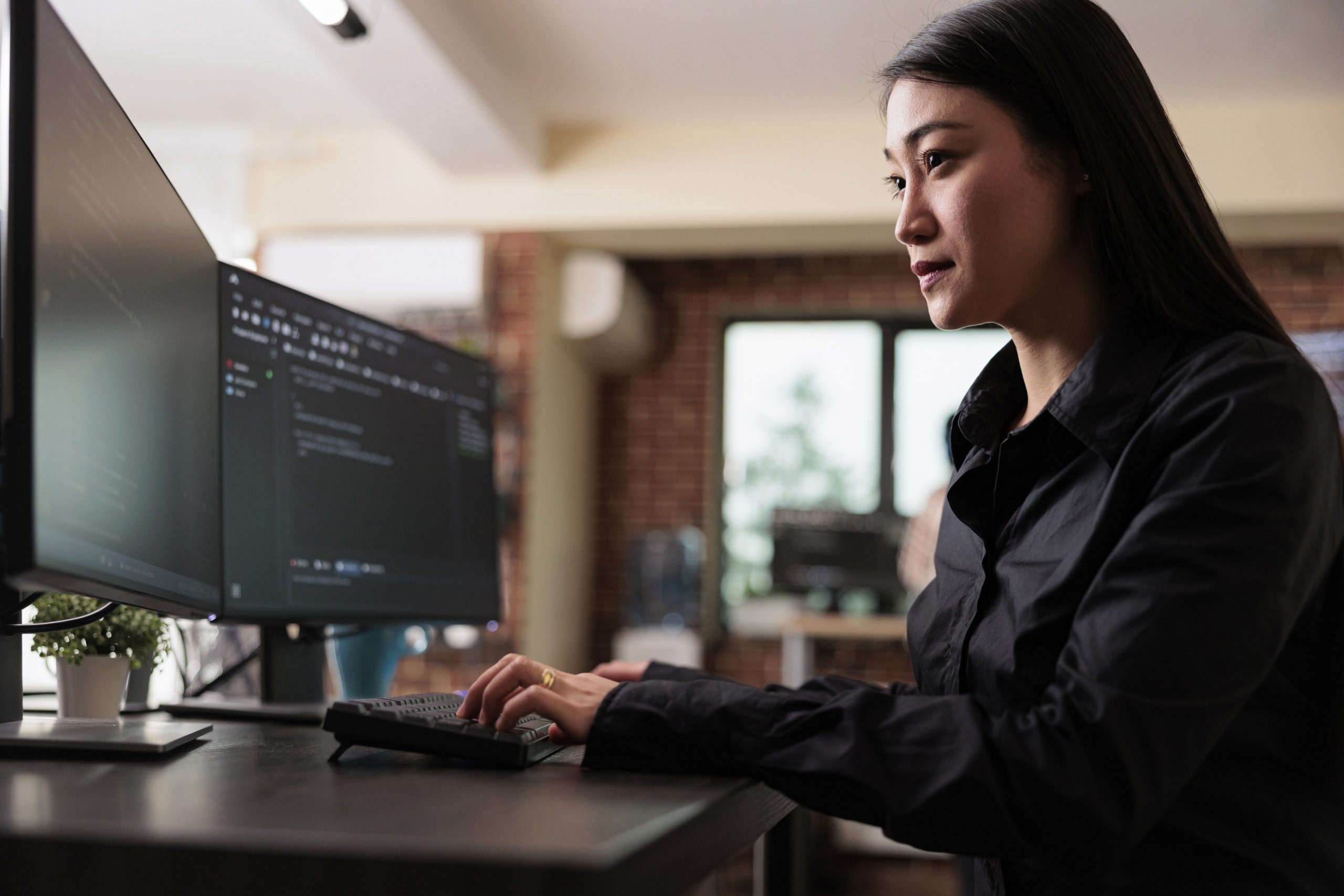 Imagem de uma mulher asiática branca de cabelos pretos cumpridos. Ela veste uma camisa preta de mangas longas e está sentada diante de uma mesa em frente a duas telas de computadores. Ela está digitando e nas telas vemos códigos de segurança.
