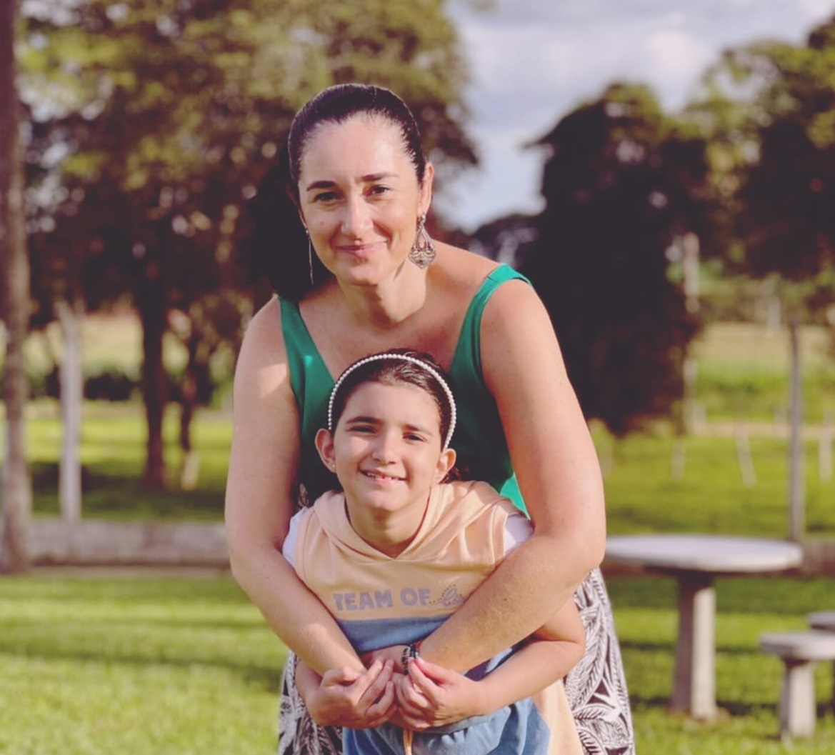 Na imagem vemos uma mulher branca de cabelos escuros presos com uma blusa verde abraçando a filha por trás. A filha é uma menina de cabelos castanhos presos. É branca. E elas estão em um campo aberto.