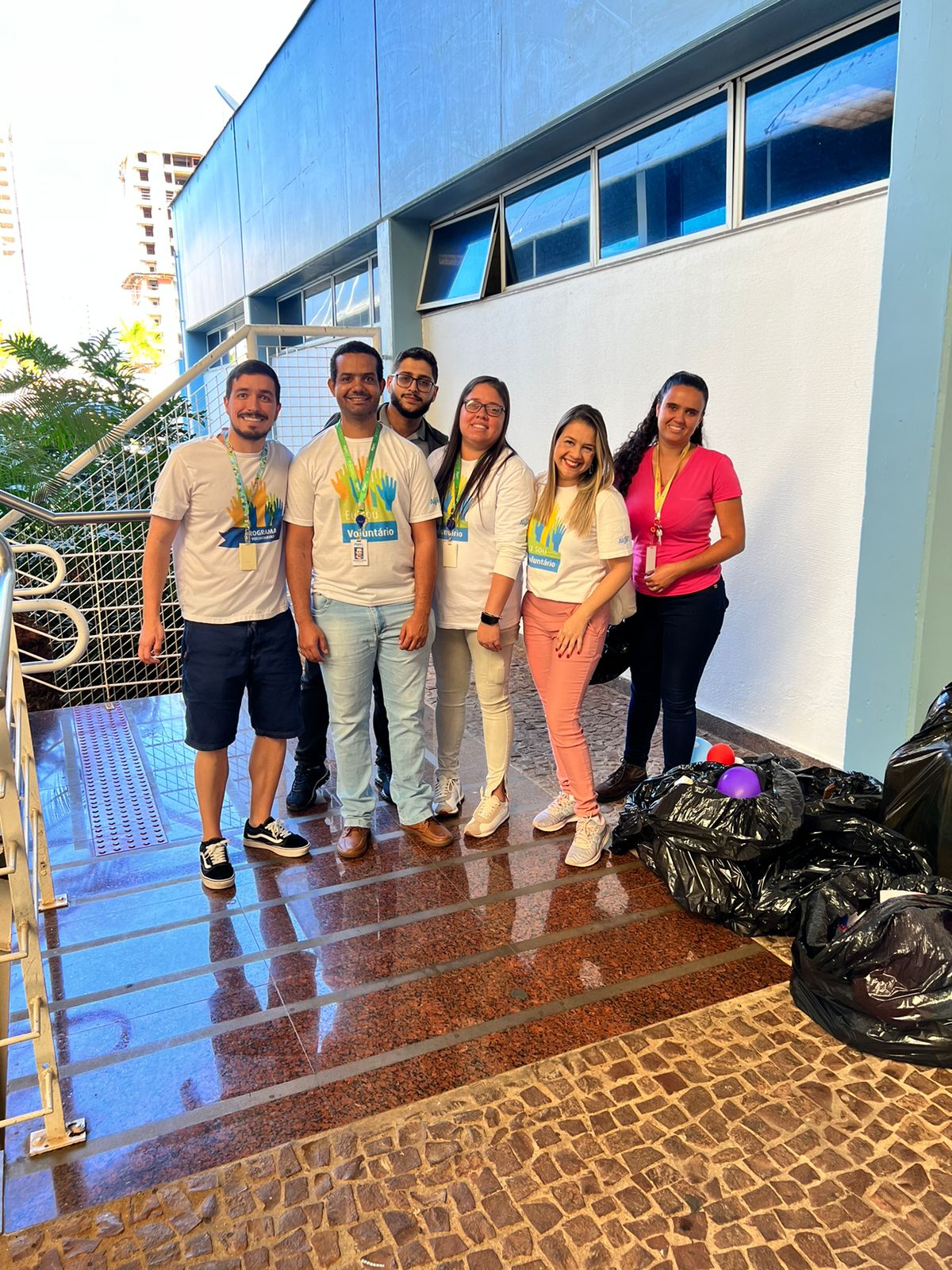 Na imagem vemos seis pessoas, três homens e três mulheres, a maioria vestindo a camisa do voluntariado. Eles estão de pé posando para foto em um local aberto, durante o dia. 