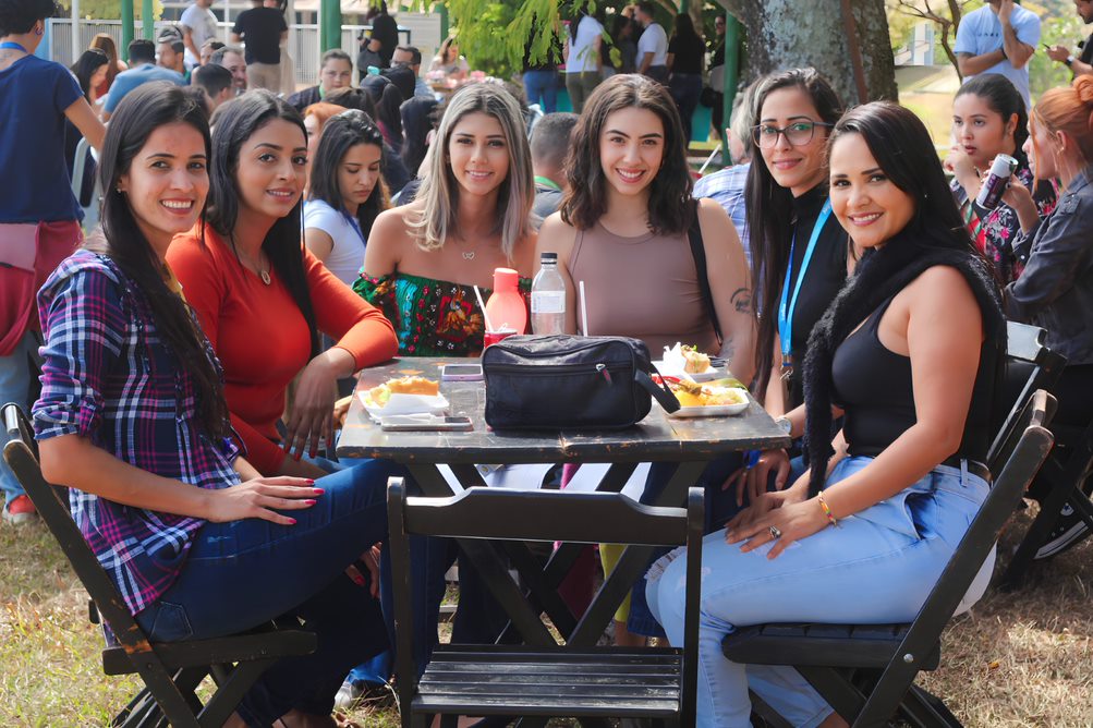 Na imagem temos um grupo de seis mulheres jovens sentadas em cadeiras de madeira escura com uma mesa no meio delas. Elas sorriem para foto. Estão em um ambiente aberto.