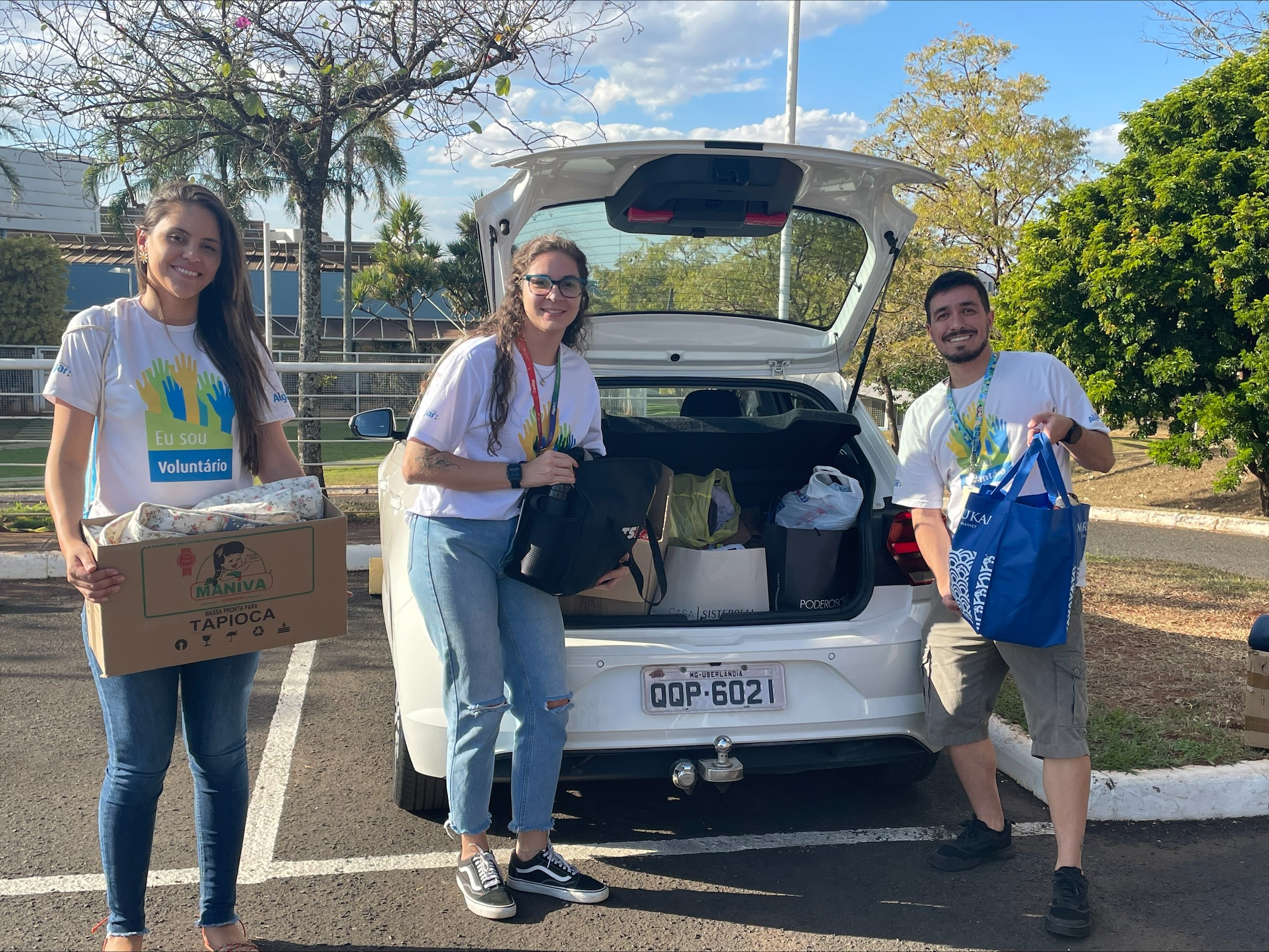 Na imagem temos duas mulheres e um homem posando para foto com a camisa do voluntariado, com doações nas mãos e um carro com o porta malas aberto.