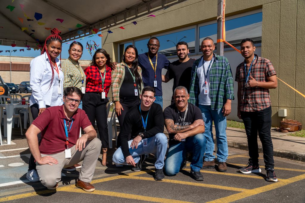 Na imagem temos um grupo de onze pessoas posando para foto. Estão em um galpão.