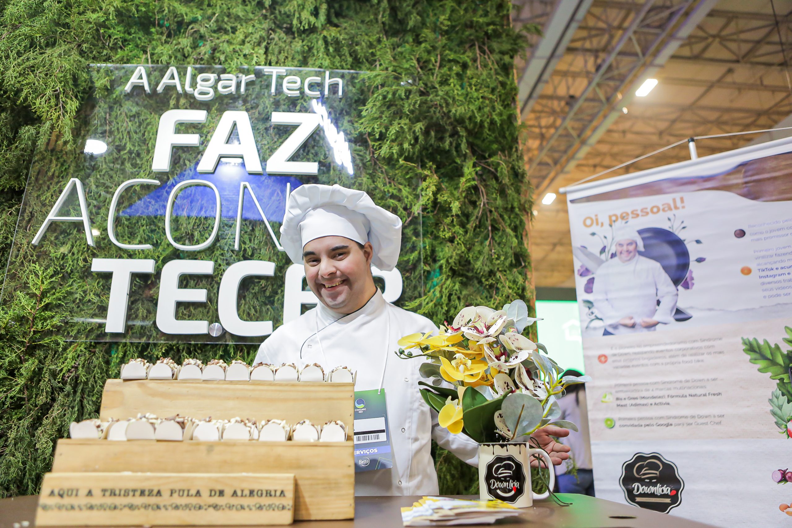 Na imagem vemos Gabriel Bernardes, um jovem branco vestindo uma doma e chapéu de confeiteiro com uma mesa de madeira com brigadeiros e atrás dele uma parede com folhagens e a frase em branco: "A Algar Tech Faz Acontecer".