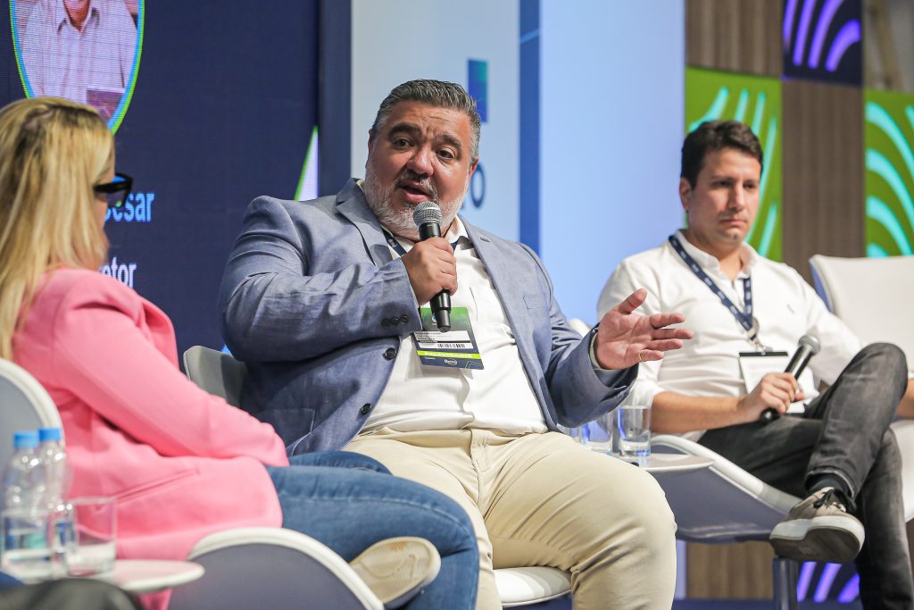 Na imagem vemos o executivo da Algar Tech Riemann Cesar usando camisa e calças brancas com blazer azul claro, sentado em uma cadeira no entre um homem e uma mulher com um microfone na mão. 