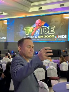 Na imagem temos o Alessandro Macedo, especialista em Cibersegurança da Algar Tech. Ele está tirandoum selfie com um telão escrito: Tande: a vida é um jogo