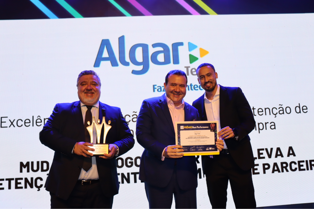 Na imagem vemos três executivos da esquerda para a direita: Homem branco, cabelos e barba grisalhos, usando terno e gravata pretos com camisa branca, segurando o troféu de bronze do Best Performance, ao lado dele temos um homem branco de cabelos pretos, usando um terno azul escuro com uma camisa clara sem gravata segurando um certificado de premiação junto com um homem alto branco, terno preto e camisa branca também sem gravata. Atrás deles temos a imagem em um telão do logo da Algar Tech e a categoria da premiação.